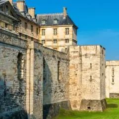 Murs imposants et tours de pierre du **château de Vincennes** sous un ciel bleu éclatant, avec des bâtiments historiques en arrière-plan.