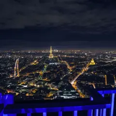 Vue nocturne de Paris depuis le rooftop de la Tour Montparnasse, avec la Tour Eiffel illuminée et un faisceau lumineux bleu traversant le ciel.