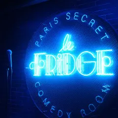 Le néon bleu "Le Fridge Comedy Club" brille sur un mur de briques. Un micro attend sur scène.