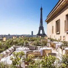 Terrasse d'un restaurant chic avec vue sur la Tour Eiffel. Tables dressées, végétation luxuriante et lanternes créent une ambiance magique.