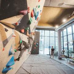 Une femme grimpe sur un mur de bloc dans un centre d'escalade moderne, le climbing district Batignolles. Un homme observe depuis le sol.