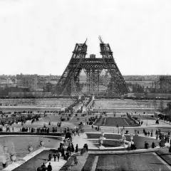 La Tour Eiffel en construction, montrant les matériaux métalliques assemblés. Des foules observent l'avancée des travaux sur le Champ de Mars.