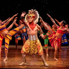 Le roi lion Mogador, en costume flamboyant, salue la foule entouré de danseurs aux couleurs vives.