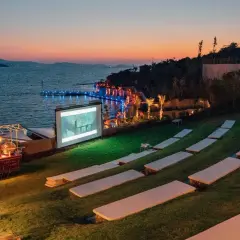 Un magnifique **cinéma plein air** au bord de la mer, avec un écran projetant un film, des sièges sur l'herbe et un chariot de pop-corn.