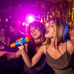 Trois amies chantent au micro lors d'une soirée karaoké à Paris. L'ambiance est festive, avec des lumières violettes et des guirlandes scintillantes.