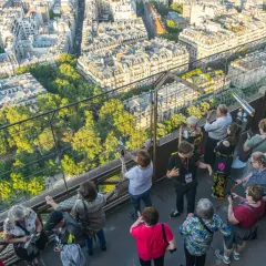 Vue panoramique de Paris depuis la Tour Eiffel, avec des touristes admirant le paysage. Le soleil d'été suggère le meilleur moment pour visiter la Tour Eiffel.