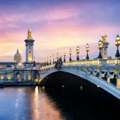 Le **pont Alexandre III** s'illumine sous un ciel crépusculaire aux teintes roses et violettes, reflétant sa splendeur dorée sur la Seine.