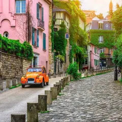 Une 2CV orange monte une rue pavée bordée de maisons roses et vertes. Parfait pour savoir que faire ce dimanche.