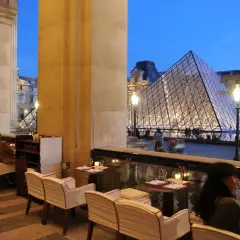 Terrasse du Café Marly, vue sur la pyramide du Louvre illuminée, avec des clients attablés.