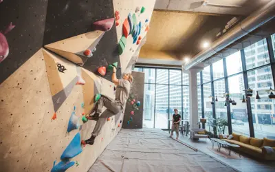 Une femme grimpe sur un mur de bloc dans un centre d'escalade moderne, le climbing district Batignolles. Un homme observe depuis le sol.