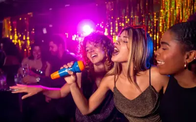 Trois amies chantent au micro lors d'une soirée karaoké à Paris. L'ambiance est festive, avec des lumières violettes et des guirlandes scintillantes.