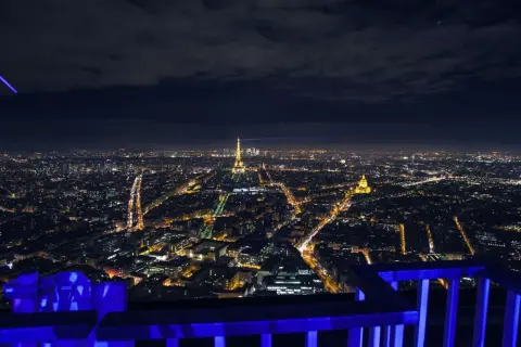 Vue nocturne de Paris depuis le rooftop de la Tour Montparnasse, avec la Tour Eiffel illuminée et un faisceau lumineux bleu traversant le ciel.