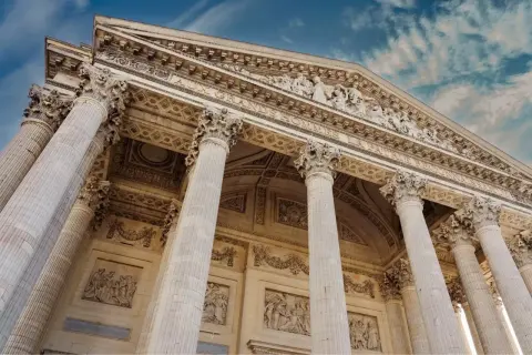 Le **Panthéon de Paris**, majestueux avec ses colonnes imposantes et ses bas-reliefs, se dresse sous un ciel bleu nuageux.