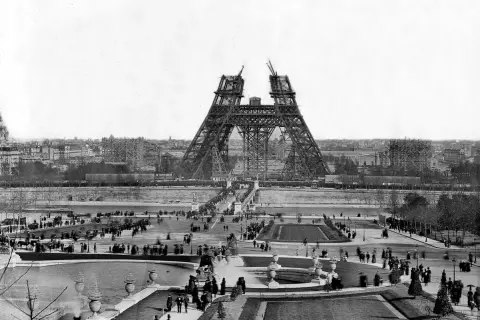 La Tour Eiffel en construction, montrant les matériaux métalliques assemblés. Des foules observent l'avancée des travaux sur le Champ de Mars.