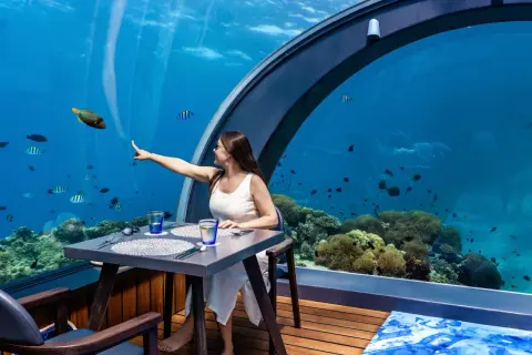 Une femme pointe un poisson dans un restaurant sous la mer, entourée de coraux et de vie marine.