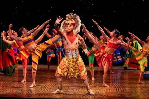 Le roi lion Mogador, en costume flamboyant, salue la foule entouré de danseurs aux couleurs vives.