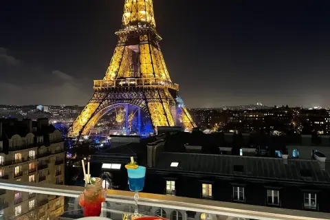 Vue nocturne de Paris depuis un balcon avec des cocktails et des tapas, la Tour Eiffel illuminée en arrière-plan. C'est le 10ème ciel !