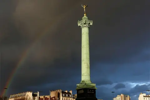 La colonne de Juillet se dresse fièrement sur la place de la Bastille, sous un ciel orageux traversé par un arc-en-ciel.