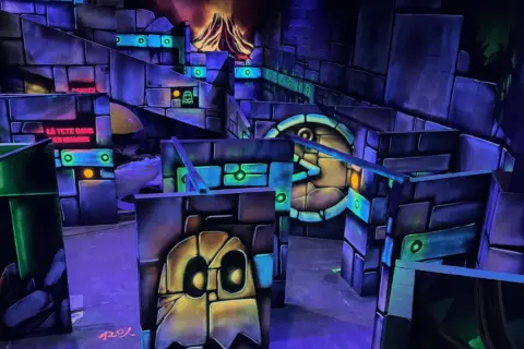 Labyrinthe de laser game aux couleurs vives, avec des fantômes et un volcan en arrière-plan. Le panneau "LA TÊTE DANS LES NUAGES" suggère une expérience amusante.