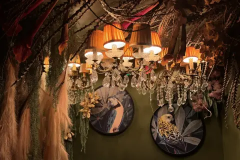 Décor luxuriant et excentrique d'un restaurant insolite à Paris, avec des lustres ornés, des plantes suspendues et des tableaux d'oiseaux exotiques.