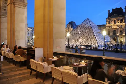 Terrasse du Café Marly, vue sur la pyramide du Louvre illuminée, avec des clients attablés.