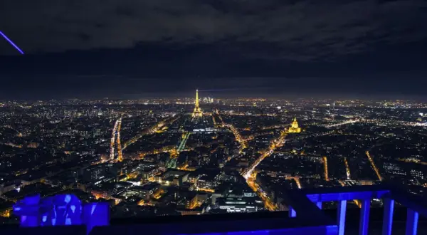 Vue nocturne de Paris depuis le rooftop de la Tour Montparnasse, avec la Tour Eiffel illuminée et un faisceau lumineux bleu traversant le ciel.