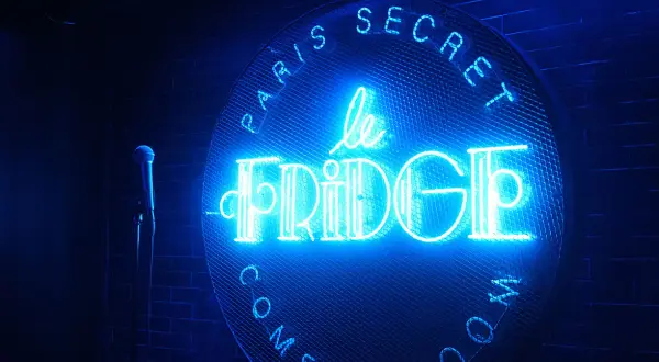 Le néon bleu "Le Fridge Comedy Club" brille sur un mur de briques. Un micro attend sur scène.