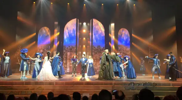 Scène du spectacle "Notre-Dame de Paris" avec des costumes d'époque et un décor évoquant la ville.