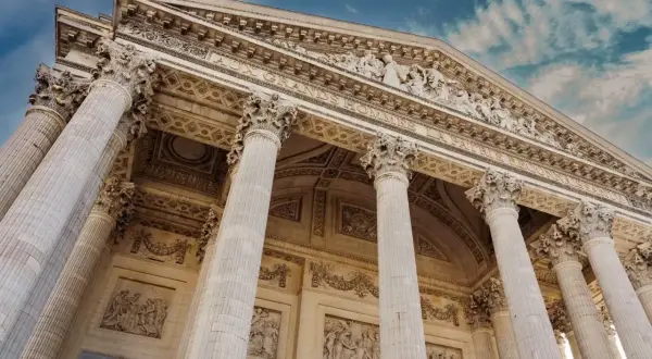 Le **Panthéon de Paris**, majestueux avec ses colonnes imposantes et ses bas-reliefs, se dresse sous un ciel bleu nuageux.