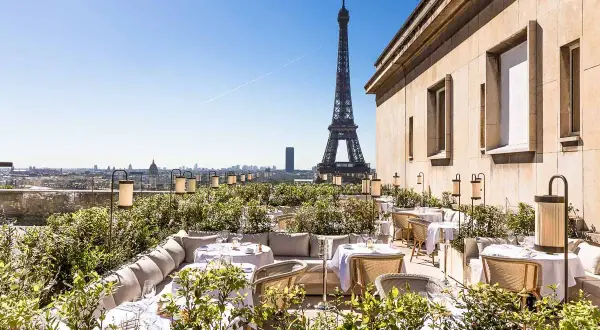 Terrasse d'un restaurant chic avec vue sur la Tour Eiffel. Tables dressées, végétation luxuriante et lanternes créent une ambiance magique.
