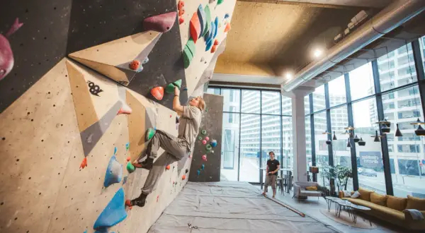 Une femme grimpe sur un mur de bloc dans un centre d'escalade moderne, le climbing district Batignolles. Un homme observe depuis le sol.