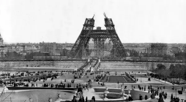 La Tour Eiffel en construction, montrant les matériaux métalliques assemblés. Des foules observent l'avancée des travaux sur le Champ de Mars.