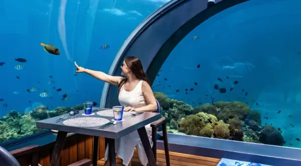 Une femme pointe un poisson dans un restaurant sous la mer, entourée de coraux et de vie marine.