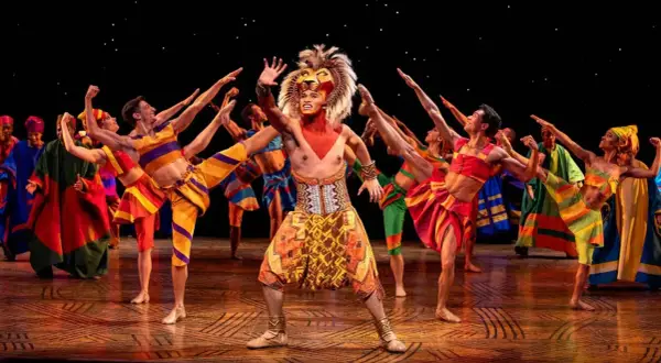 Le roi lion Mogador, en costume flamboyant, salue la foule entouré de danseurs aux couleurs vives.