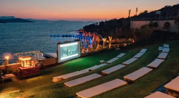 Un magnifique **cinéma plein air** au bord de la mer, avec un écran projetant un film, des sièges sur l'herbe et un chariot de pop-corn.
