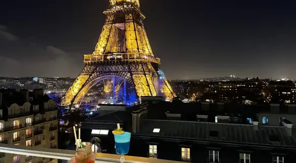 Vue nocturne de Paris depuis un balcon avec des cocktails et des tapas, la Tour Eiffel illuminée en arrière-plan. C'est le 10ème ciel !