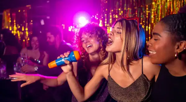 Trois amies chantent au micro lors d'une soirée karaoké à Paris. L'ambiance est festive, avec des lumières violettes et des guirlandes scintillantes.