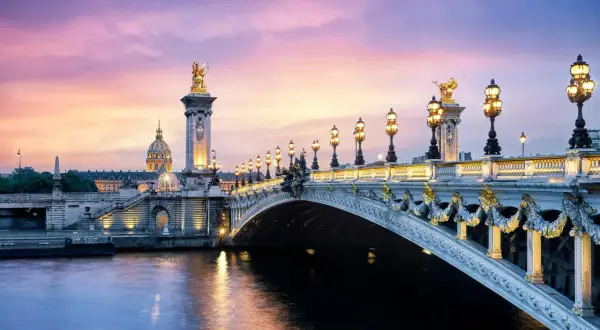 Le **pont Alexandre III** s'illumine sous un ciel crépusculaire aux teintes roses et violettes, reflétant sa splendeur dorée sur la Seine.