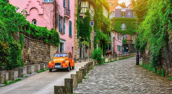 Une 2CV orange monte une rue pavée bordée de maisons roses et vertes. Parfait pour savoir que faire ce dimanche.
