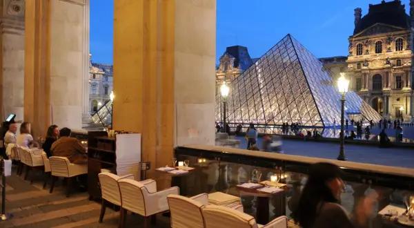 Terrasse du Café Marly, vue sur la pyramide du Louvre illuminée, avec des clients attablés.