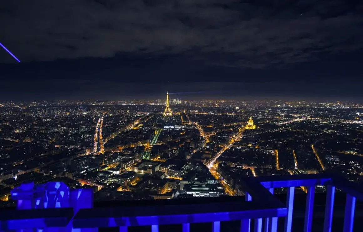 Vue nocturne de Paris depuis le rooftop de la Tour Montparnasse, avec la Tour Eiffel illuminée et un faisceau lumineux bleu traversant le ciel.