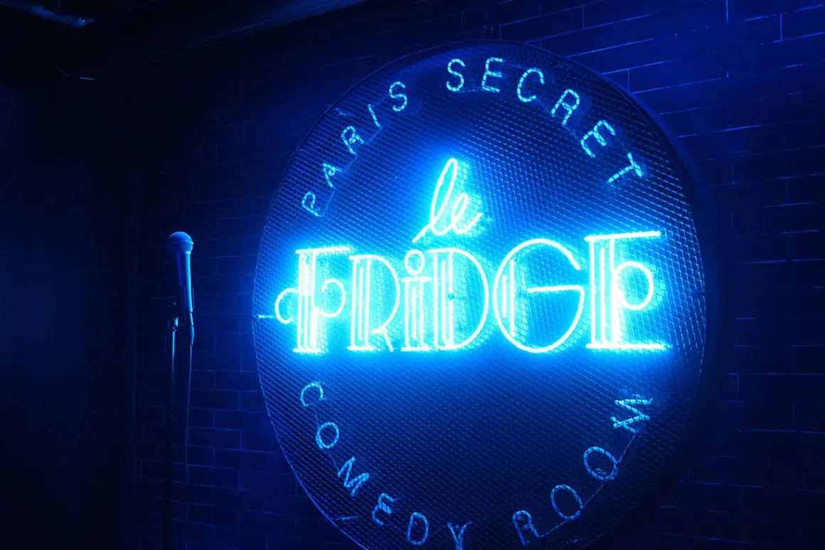 Le néon bleu "Le Fridge Comedy Club" brille sur un mur de briques. Un micro attend sur scène.