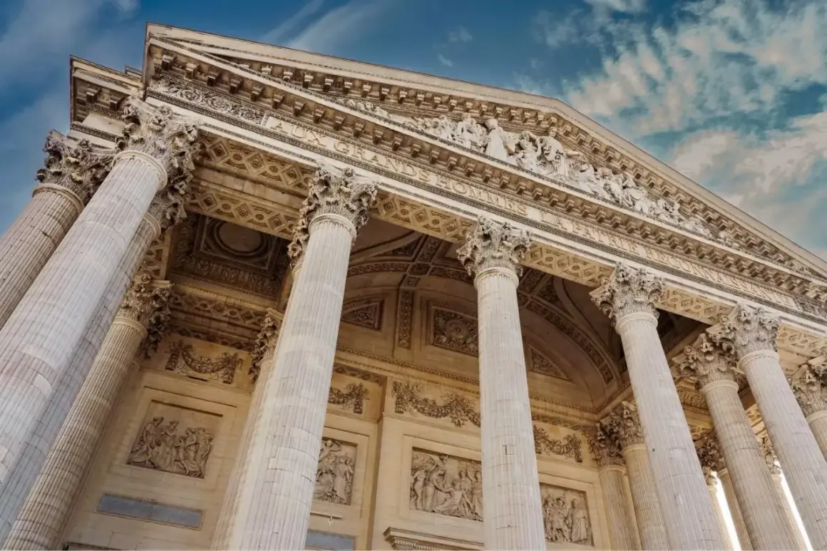 Le **Panthéon de Paris**, majestueux avec ses colonnes imposantes et ses bas-reliefs, se dresse sous un ciel bleu nuageux.