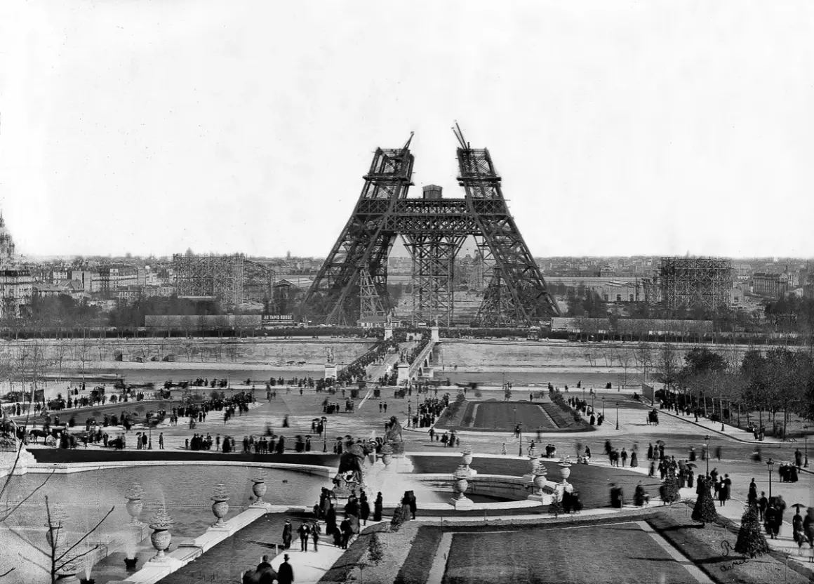 La Tour Eiffel en construction, montrant les matériaux métalliques assemblés. Des foules observent l'avancée des travaux sur le Champ de Mars.