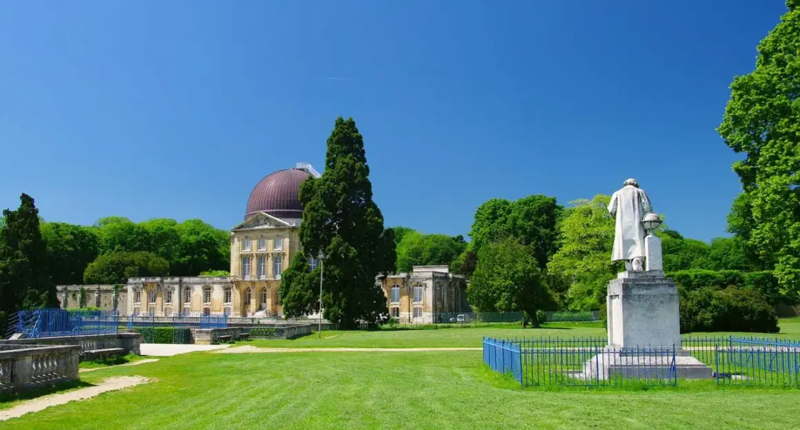 Un grand bâtiment avec un dôme rouge, entouré d'arbres verts et d'une pelouse. Une statue se dresse devant, comme si elle surveillait le hangar Y.