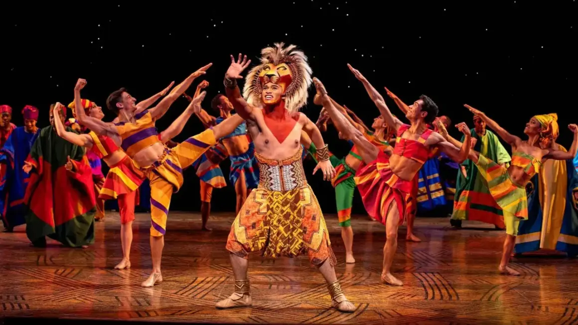 Le roi lion Mogador, en costume flamboyant, salue la foule entouré de danseurs aux couleurs vives.