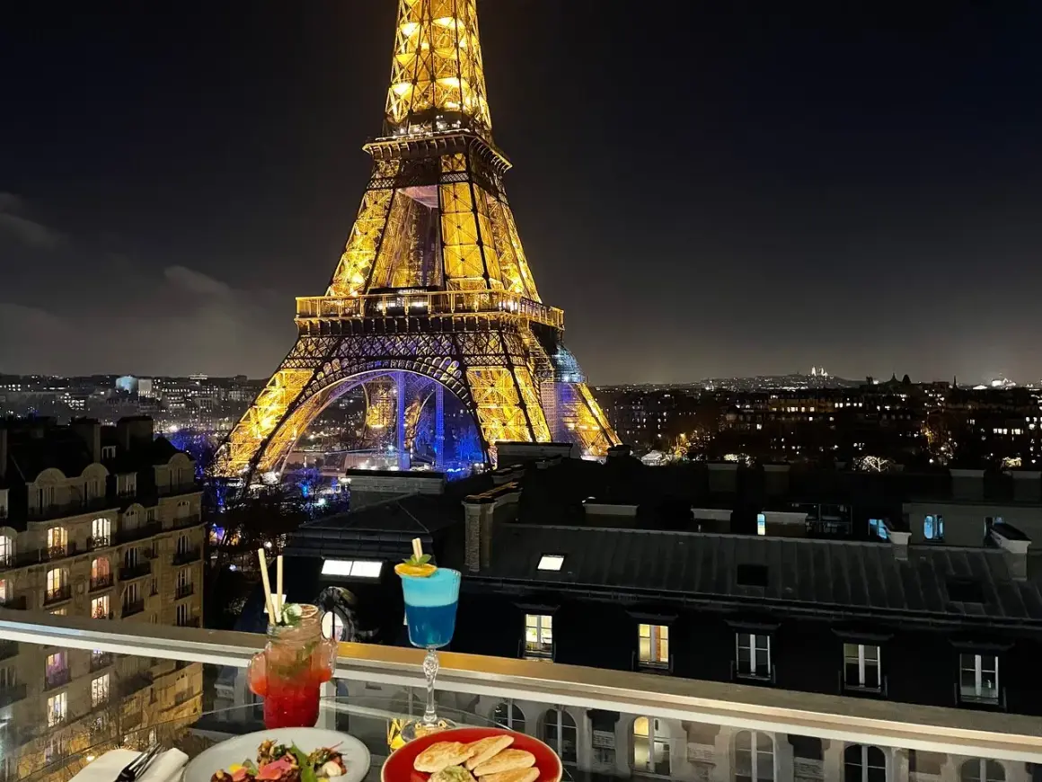Vue nocturne de Paris depuis un balcon avec des cocktails et des tapas, la Tour Eiffel illuminée en arrière-plan. C'est le 10ème ciel !