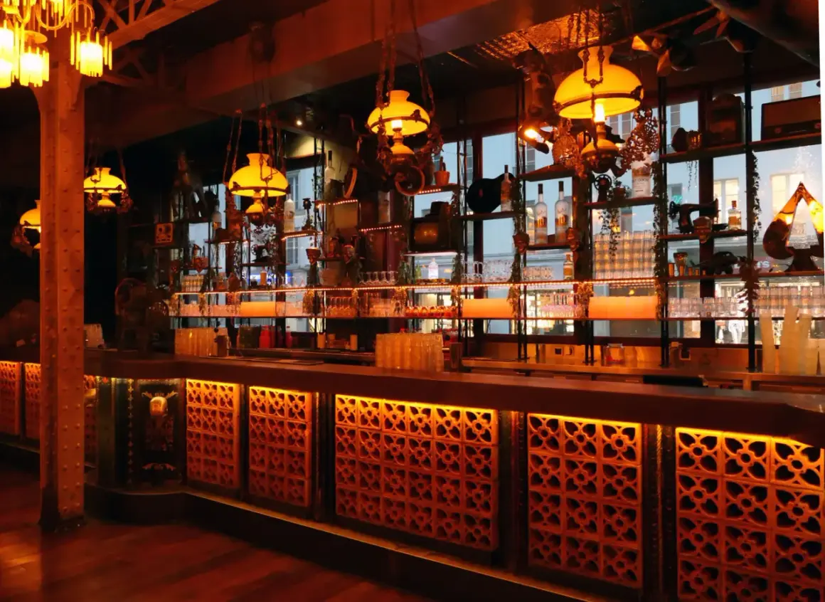 Ambiance chaleureuse d'un bar parisien, avec des lumières tamisées, des étagères remplies de bouteilles et de bibelots, évoquant un **barrio latino** à Paris.