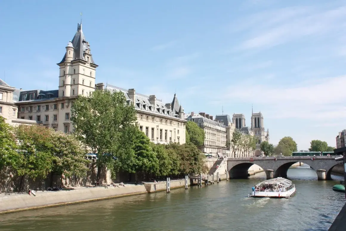 Bateau de croisière naviguant sur la Seine, passant sous un pont. Le quai de la photo est bordé d'arbres et de bâtiments parisiens.