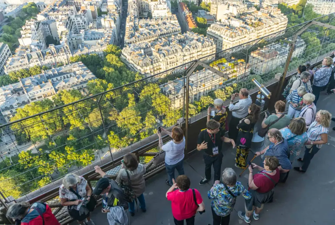 Vue panoramique de Paris depuis la Tour Eiffel, avec des touristes admirant le paysage. Le soleil d'été suggère le meilleur moment pour visiter la Tour Eiffel.